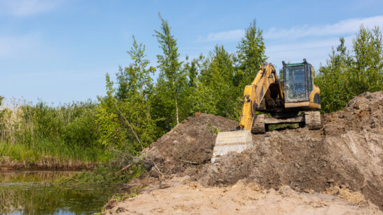 A yellow tracked excavator works along a wetland pond bank, moving soil and clearing vegetation for land development and erosion control. Bright blue sky and green trees frame the industrial scene